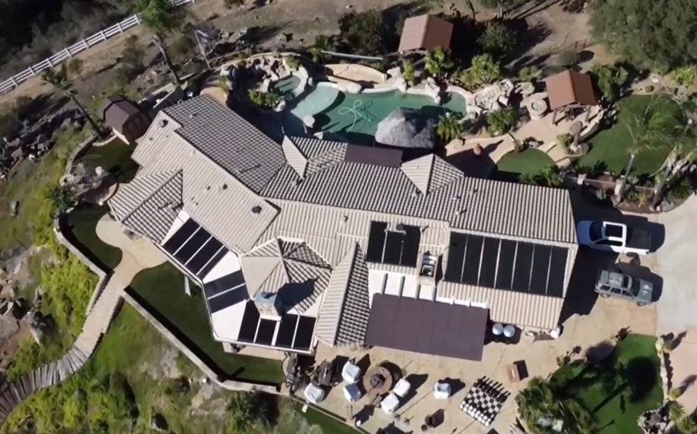 Aerial view of a tile-roof home with solar pool heating collectors on several roof planes above a backyard pool with a rock waterfall